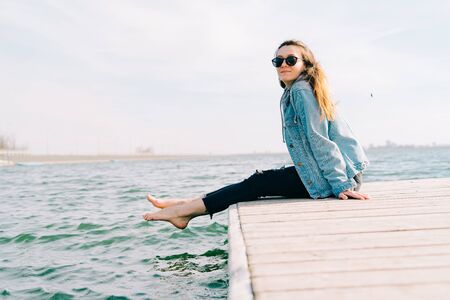 a young beautiful stylish girl is sitting on a pier near the lakeの写真素材