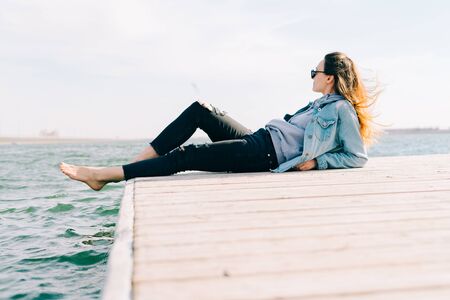 a young beautiful stylish girl is sitting on a pier near the lakeの写真素材