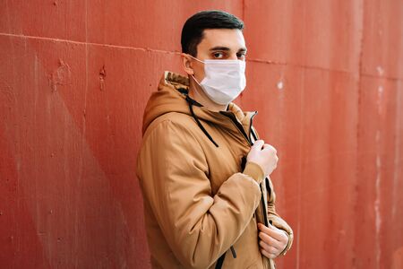 a young man in a brown jacket with a medical mask on his face on the street, near the red wall....