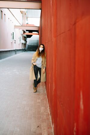 a young girl in a brown coat with a medical mask on her face on the street, near the red wall. protection from the virusの写真素材