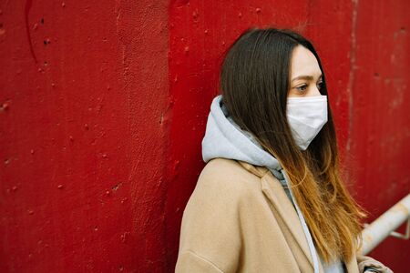 a young girl in a brown coat with a medical mask on her face on the street, near the red wall....