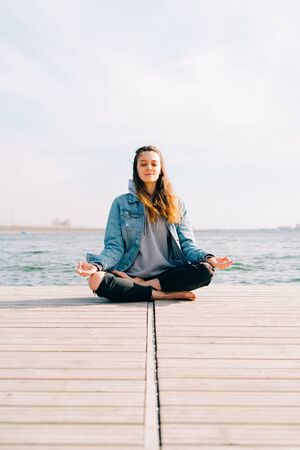 a young beautiful girl is sitting on a pier near the lake and doing meditation practiceの写真素材