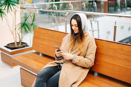 a young girl in a brown coat is sitting on a bench in a shopping center and using her smartphone. lifestyleの写真素材