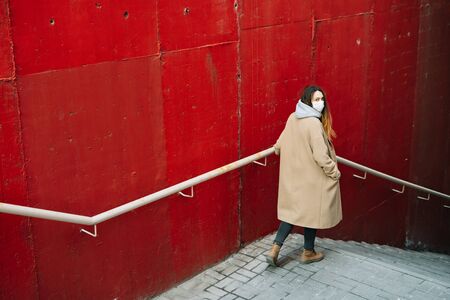 a young girl in a brown coat with a medical mask on her face on the street, near the red wall. protection from the virusの写真素材