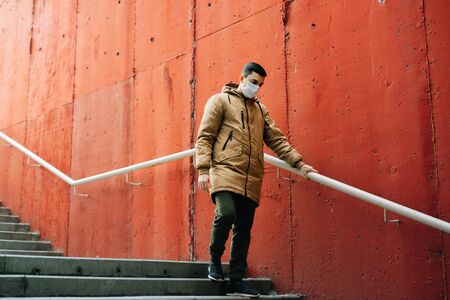 a young man in a brown jacket with a medical mask on his face on the street, near the red wall....