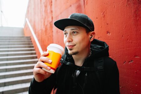 a young guy in black sportswear and a baseball cap on the street, standing near a red wall and drinking coffee .の写真素材
