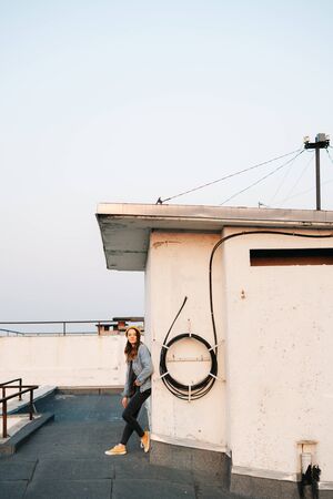 white European woman stands near the wall on the roof of a multi-storey building in stylish denim clothing and a trendy yellow hat .in the rays of the setting sun.lifestyle concept. fashion seasonsの写真素材