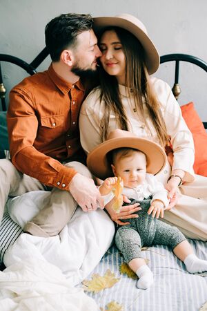 a happy stylish family of three in trendy shades of clothing, sitting on the bed, mom and dad hugging their little son and smiling at each other in a spacious Studioの写真素材