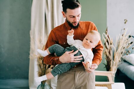 a young stylish father in trend orange shirt plays with his young son in a brown hat in his arms in...