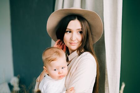 young mother in trend beige dress and hat, holding her stylishly dressed son in her arms in a spacious Studio.の写真素材
