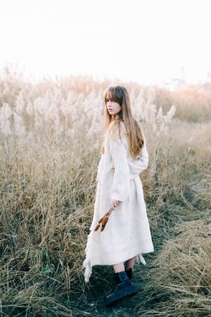 portrait of a young beautiful girl in a white dress, a warm knitted cardigan and black shoes among dry fluffy reedsの写真素材