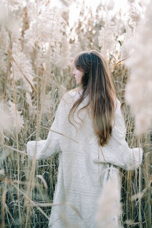 portrait of a young beautiful girl in a white dress, a warm knitted cardigan and black shoes among dry fluffy reedsの写真素材