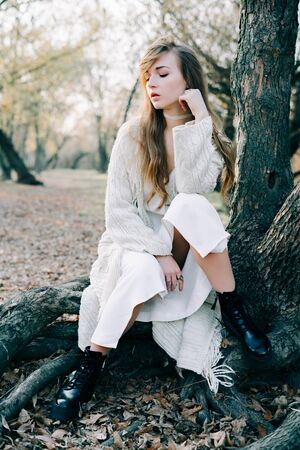 portrait of a young attractive blonde girl in a light white dress and black shoes stands on the root of a treeの写真素材