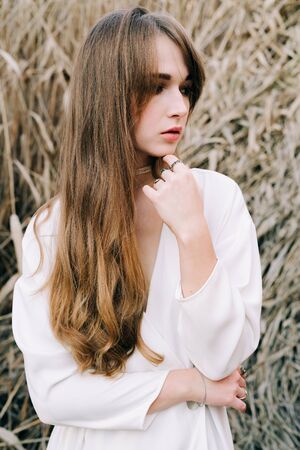 portrait of a young attractive girl in a light white dress and black shoes standing among the reedsの写真素材