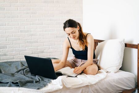European woman, using a laptop, studying information on the Internet in her bed, writes notes in her diary,while the virus is quarantined.Cozy workplace, e-learning conceptの写真素材