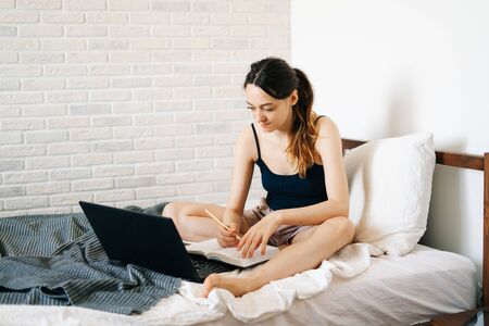 European woman, using a laptop, studying information on the Internet in her bed, writes notes in her diary,while the virus is quarantined.Cozy workplace, e-learning conceptの写真素材