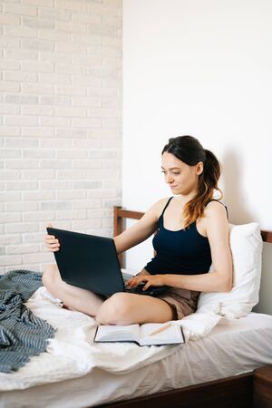 A young woman, using a laptop, communicates over the Internet with a client in her bed, while the virus is quarantined.Cozy office workplace, Remote work, e-learning conceptの写真素材
