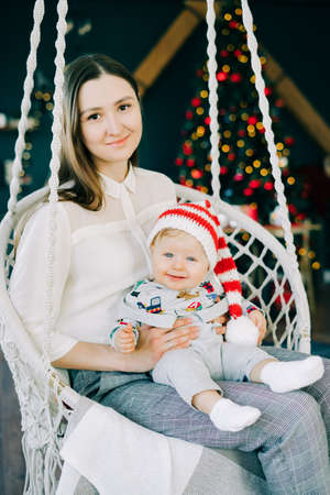the child's first Christmas.mom holds with a little boy in a Santa hat, sitting in a wicker swing on the background of a bright glowing Christmas tree Christmas concept.の写真素材