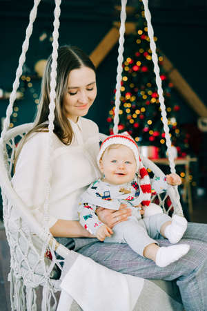 the child's first Christmas.mom holds with a little boy in a Santa hat, sitting in a wicker swing on the background of a bright glowing Christmas tree Christmas concept.の写真素材
