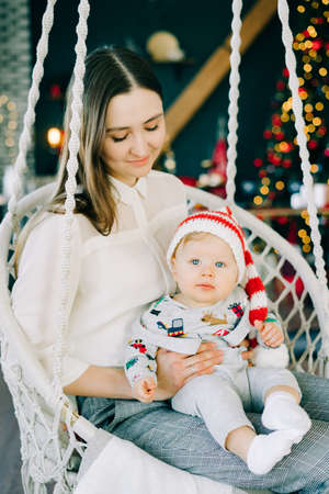 the child's first Christmas.mom holds with a little boy in a Santa hat, sitting in a wicker swing on the background of a bright glowing Christmas tree Christmas concept.の写真素材