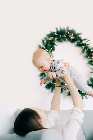 the child's first Christmas.mom lies on the bed and holds a little boy in her arms against the background of a white wall with a wreath of Christmas trees.の写真素材