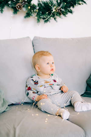 the child's first Christmas.a cute little boy is sitting in a festive costume on a gray sofa, against a white wall with a natural wreath of spruce. Christmas concept.の写真素材