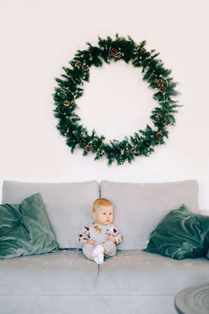 the child's first Christmas.a cute little boy is sitting in a festive costume on a gray sofa, against a white wall with a natural wreath of spruce. Christmas concept.の写真素材