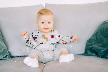 a small cute, cheerful boy sits in a festive Christmas costume on a gray sofa and plays with a bright light garland.Christmas concept.の写真素材