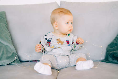 a cute little boy who is teething is sitting in a festive Christmas costume on a gray sofa and playing with a bright light garland.Christmas concept.の写真素材