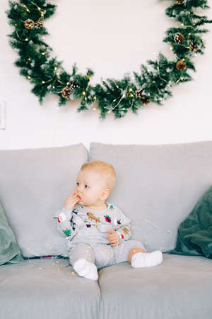 the child's first Christmas.a cute little boy is sitting in a festive costume on a gray sofa, against a white wall with a natural wreath of spruce. Christmas concept.の写真素材