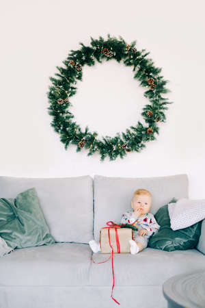 the child's first Christmas. a cute little boy is sitting in a festive costume on a gray sofa with a large gift with a red ribbon, against a white wall with a natural fir wreath. Christmas concept.の写真素材