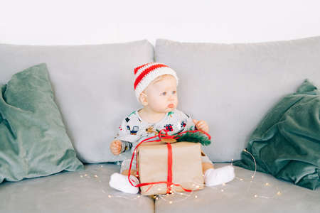 brooding little boy sits in Santa hat and holiday costume on gray sofa with a large gift with red ribbon, against white wall with natural fir wreath.Christmas concept.の写真素材