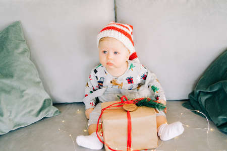brooding little boy sits in Santa hat and holiday costume on gray sofa with a large gift with red ribbon, against white wall with natural fir wreath.Christmas concept.の写真素材