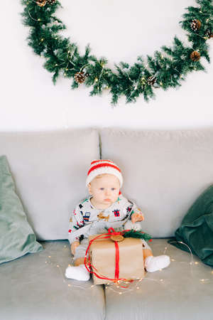 brooding little boy sits in Santa hat and holiday costume on gray sofa with a large gift with red...