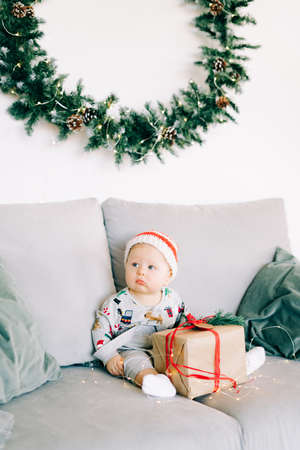 brooding little boy sits in Santa hat and holiday costume on gray sofa with a large gift with red ribbon, against white wall with natural fir wreath.Christmas concept.の写真素材