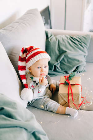 brooding little boy sits in Santa hat and holiday costume on gray sofa with a large gift with red ribbon, against white wall with natural fir wreath.Christmas concept.の写真素材