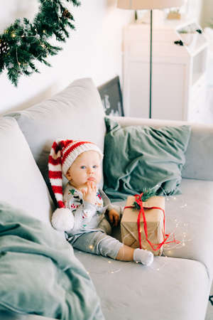 brooding little boy sits in Santa hat and holiday costume on gray sofa with a large gift with red...