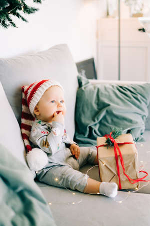 brooding little boy sits in Santa hat and holiday costume on gray sofa with a large gift with red ribbon, against white wall with natural fir wreath.Christmas concept.の写真素材