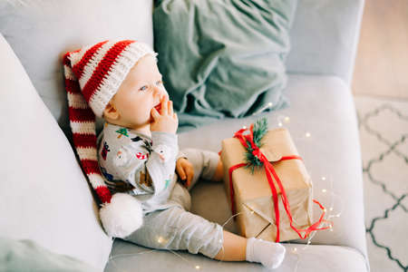 brooding little boy sits in Santa hat and holiday costume on gray sofa with a large gift with red ribbon, against white wall with natural fir wreath.Christmas concept.の写真素材
