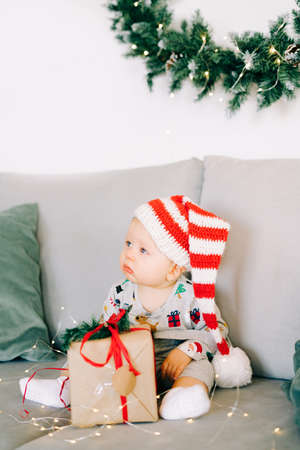 brooding little boy sits in Santa hat and holiday costume on gray sofa with a large gift with red ribbon, against white wall with natural fir wreath.Christmas concept.の写真素材