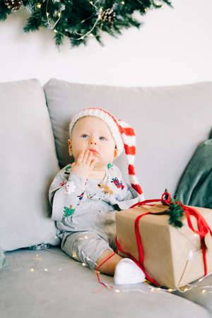 brooding little boy sits in Santa hat and holiday costume on gray sofa with a large gift with red ribbon, against white wall with natural fir wreath.Christmas concept.の写真素材