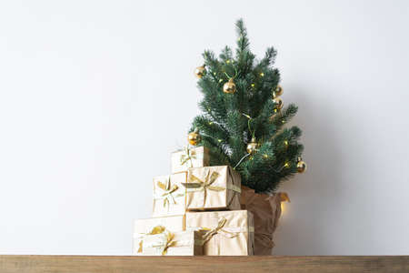 a small Christmas tree with warm bulbs in a pot on a wooden table with gifts wrapped in gold paper. the concept of Christmasの写真素材