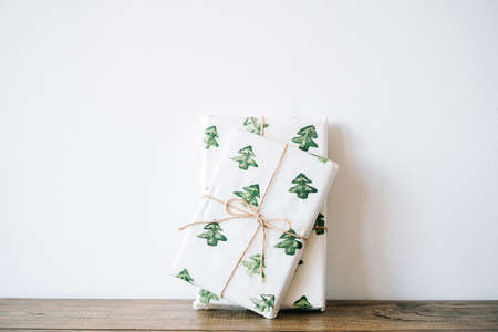 the New Year's gift is packed in festive paper with watercolor Christmas trees on a wooden table against a white wall. High quality photoの写真素材