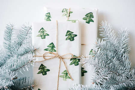the New Year's gift is packed in festive paper with watercolor Christmas trees on a wooden table against a white wall with white spruce branches. High quality photoの写真素材