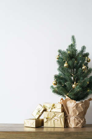 a small Christmas tree with warm bulbs in a pot on a wooden table with gifts wrapped in gold paper. the concept of Christmasの写真素材