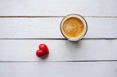 Glass of coffee on white wooden table. Top view. Red heart is near the glass of coffee. Copy space.の写真素材