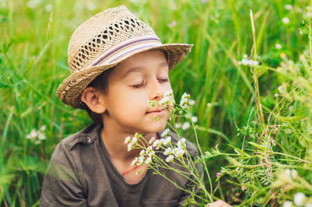 Adorable kid boy in straw hat sitting on a summer meadow and smelling white flowers. Sunset in the park. Outdoors. Vacations. Walking in the farm.の写真素材