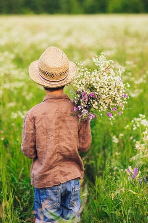 Adorable kid boy in straw hat sitting on a summer meadow and holding flowers  in his hands. Sunset in the park. Outdoors. Vacations. Walking in the farm.の写真素材