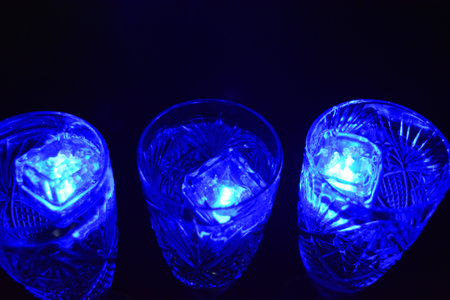 Crystal champagne glasses with carvings in the form of snowflakes are located on a black matte background. Bright multicolor doidic glowing ice cubes float in the drink.の写真素材