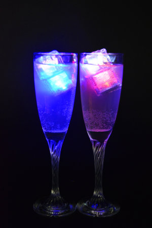A champagne glass on a long swirling legs with a white drink is located on a black background. Luminous ice cubes of different colors with led diode illumination float in a glass.の写真素材